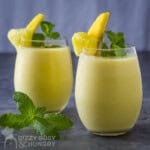 Side shot of two cocktail glasses with pineapple smoothie garnished with mango and mint with another mint garnish on the side.