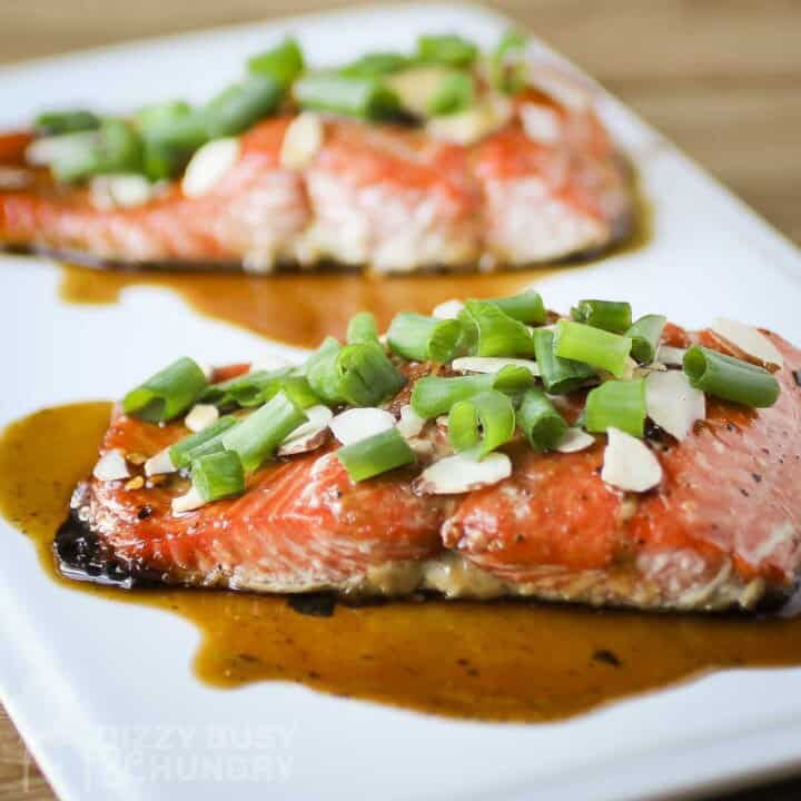 Side shot of two pieces of salmon garnished with green onions, almonds, and sauce on a white square plate.