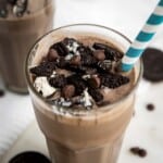 Angled close up shot of an oreo milkshake garnished with oreos and chocolate chips with a blue and white striped straw.