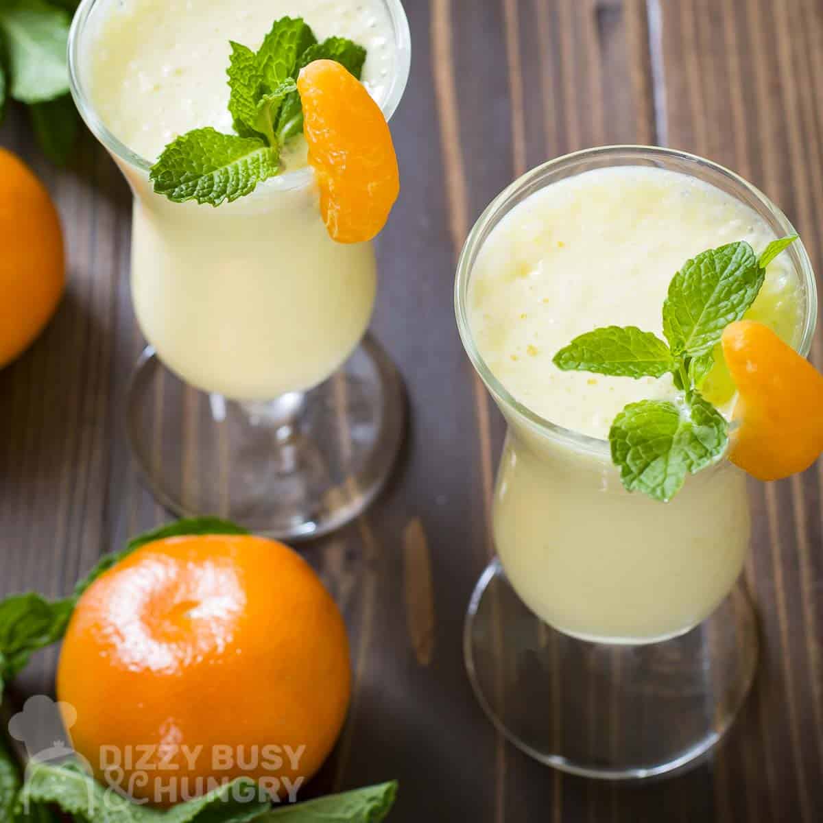 Overhead angled shot of two cocktail glasses with orange pineapple smoothie garnished with orange slices and mint with whole oranges and mint sprigs on the side.