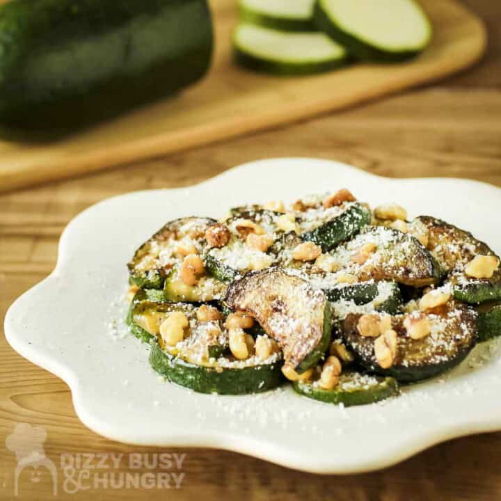 Side shot of sauteed zucchini and walnuts with parmesan cheese on a white decorative plate with raw zucchini on a cutting board in the background.