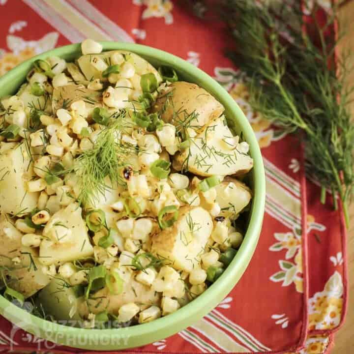 Overhead shot of potato salad with corn garnished with dill and green onions in a green bowl on a red decorative cloth with dill sprigs on the side.