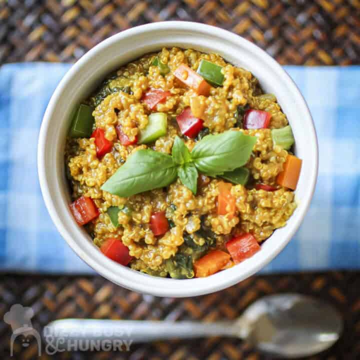 Overhead shot of quinoa curry garnished with fresh herbs in a white bowl on a blue plaid cloth with a spoon on the side.