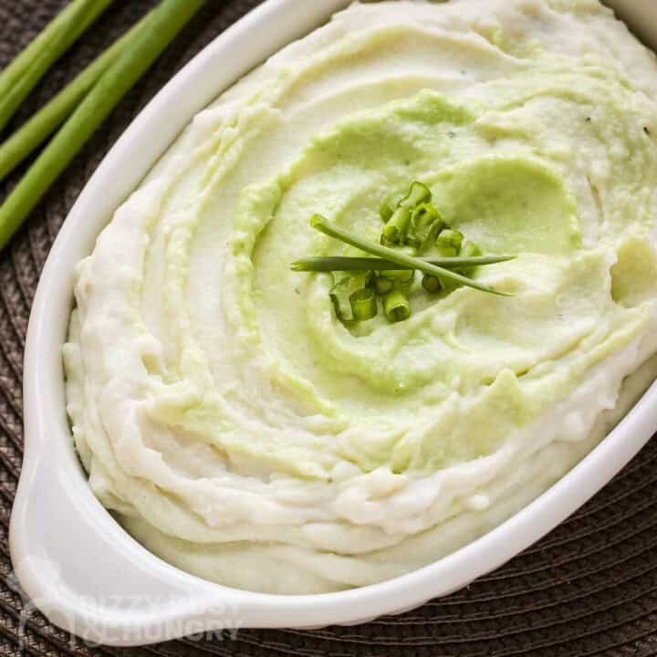 Overhead close up shot of wasabi mashed potatoes in a white baking dish garnished with green onions and chives with more green onions in the background.