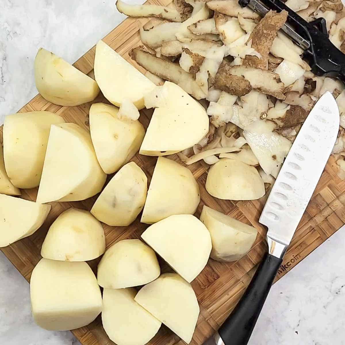 Overhead view of potatoes cut into chunks on a wooden cutting board next to a cutting knife.