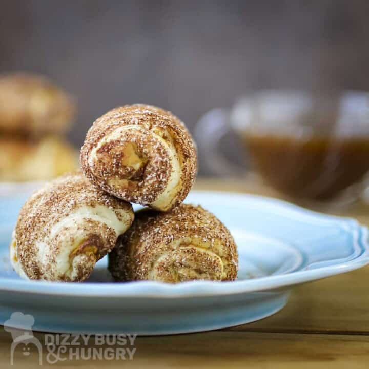 Side close up shot of three pumpkin crescent rolls stacked on a blue plate with a clear bowl of caramel in the background.