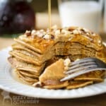 Side close up shot of pumpkin pancakes stacked on each other with a fork taking slices out and garnished with sliced almonds and maple syrup on a white plate.