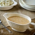 Side shot of gravy in a decorative ceramic dish with a tray of mashed potatoes and a wooden spoon in the background all on a burlap cloth.