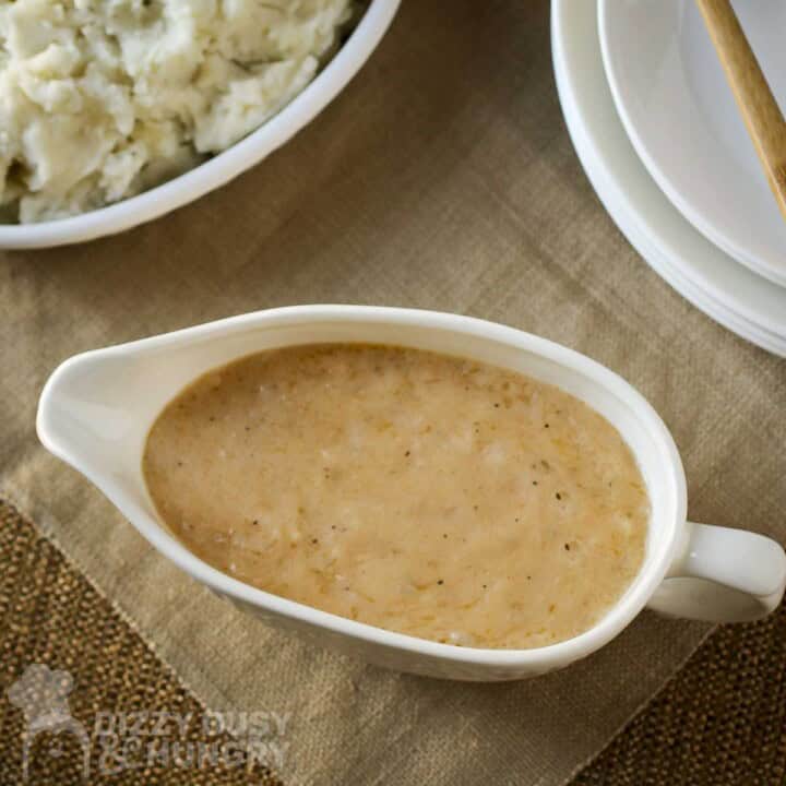 Overhead shot of gravy in a ceramic dish on a burlap cloth with mashed potatoes and more white plates on the side.