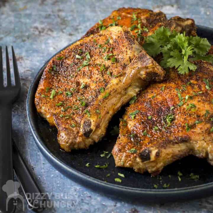 Side shot of three pork chops on a black plate garnished with fresh parsley with a black fork on the side.