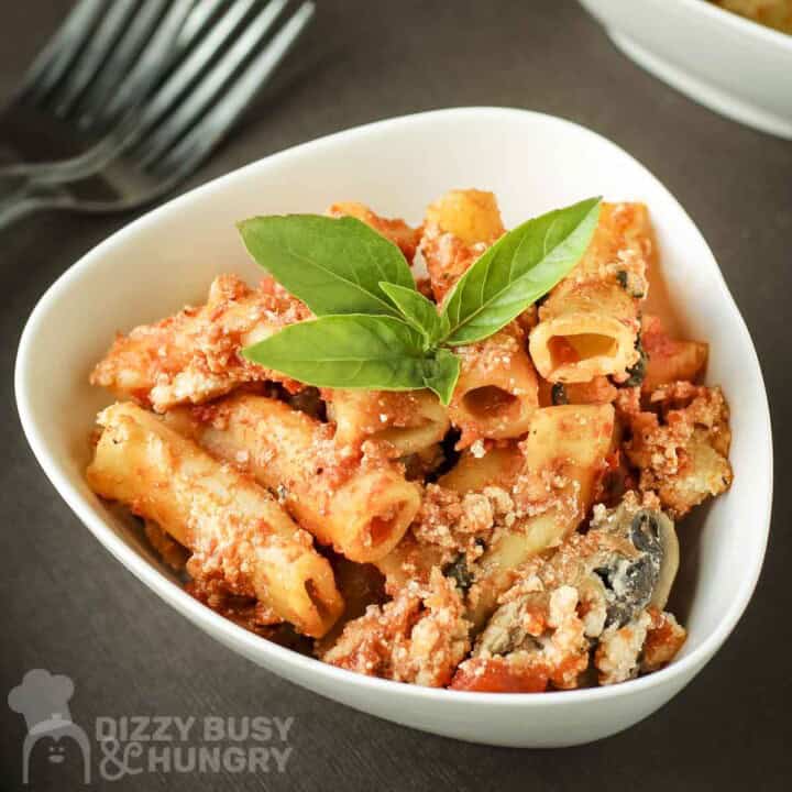 Close up shot of a white bowl with baked ziti garnished with fresh basil on a brown surface.