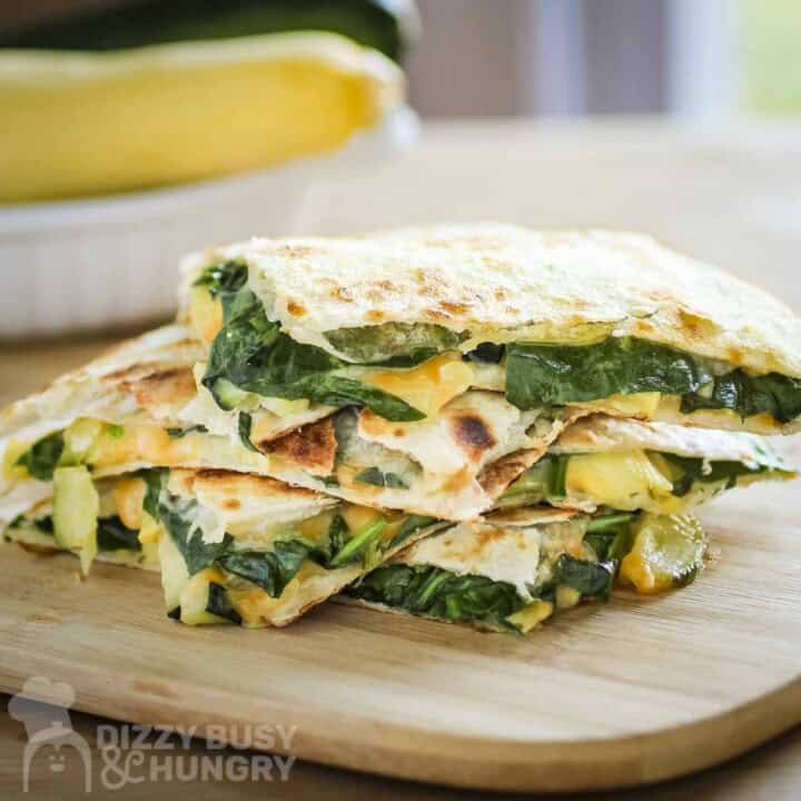 Overhead shot of three halves of spinach quesadillas stacked on a wooden cutting board with a bowl with zucchinis in the background.