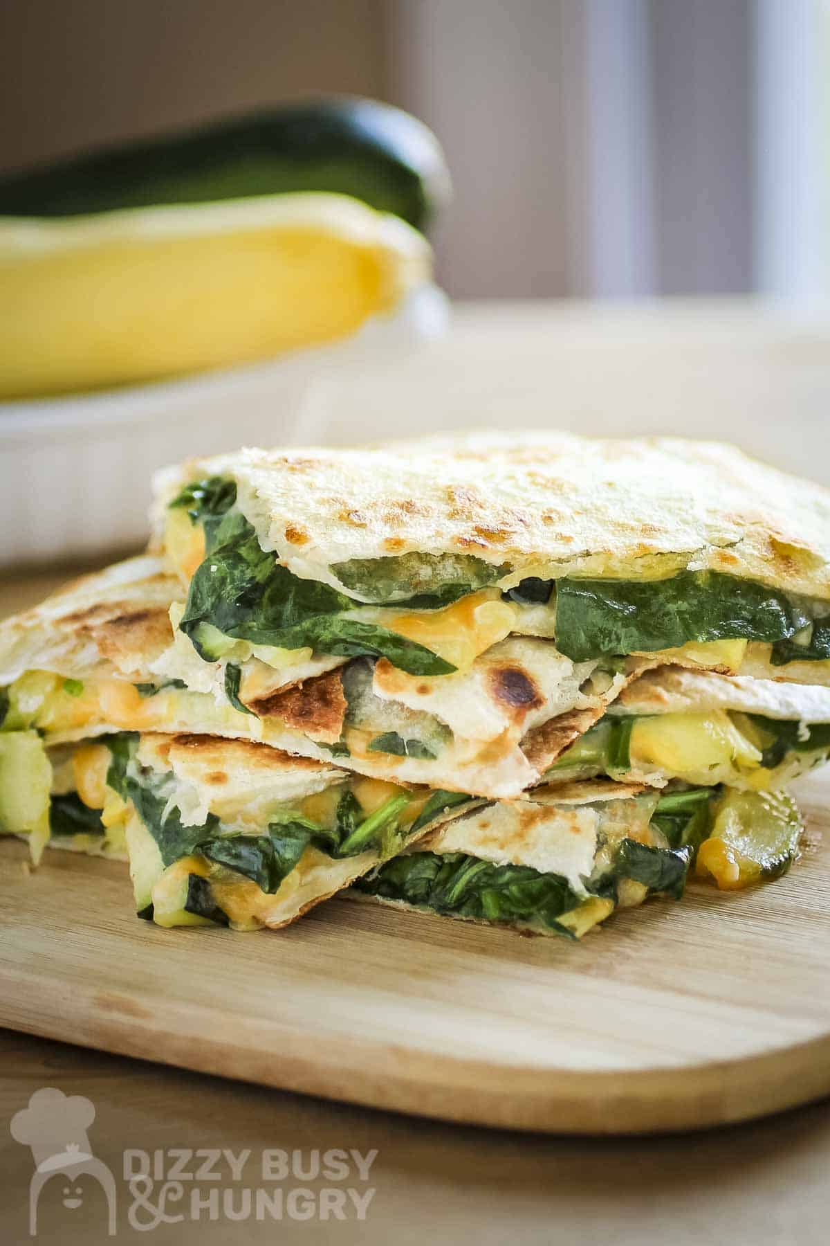 Overhead shot of three halves of spinach quesadillas stacked on a wooden cutting board with a bowl with zucchini in the background.