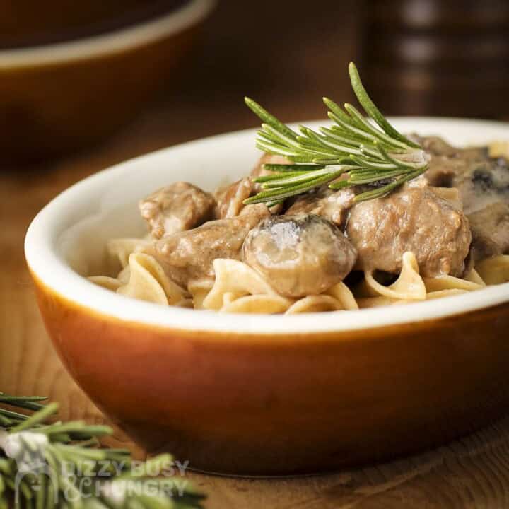 Side shot of crockpot beef stroganoff garnished with rosemary in an orange oval bowl on a wooden surface.