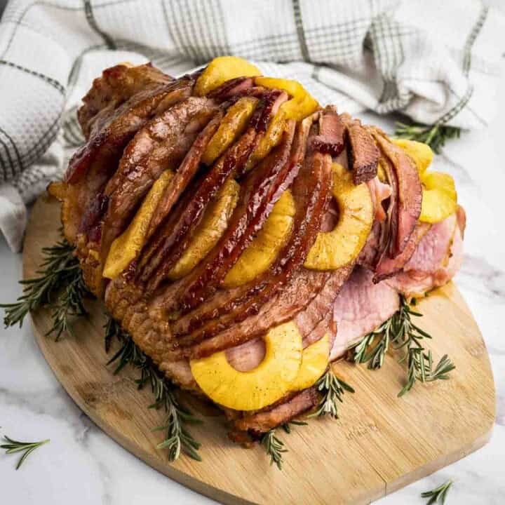 Side shot of ham with pinapple on a wooden cutting board garnished with rosemary sprigs on a marble surface.