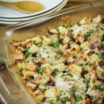 Side shot of chicken potato bake in a casserole dish on a wooden table with plates in the background.