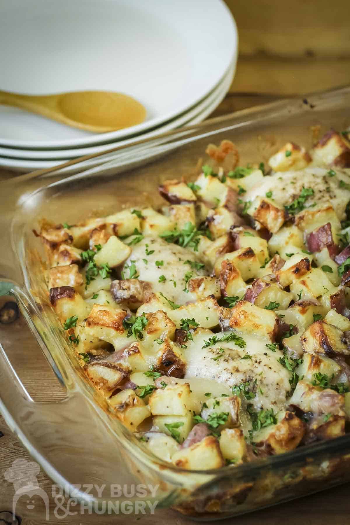 Side shot of chicken potato bake in a casserole dish on a wooden table with plates in the background.