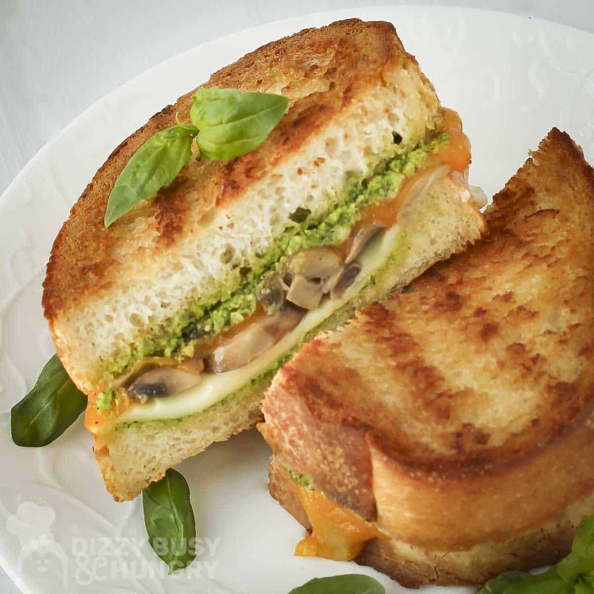 Angled shot of two halves of grilled cheese mushrooom sandwich on a white plate garnished with basil.