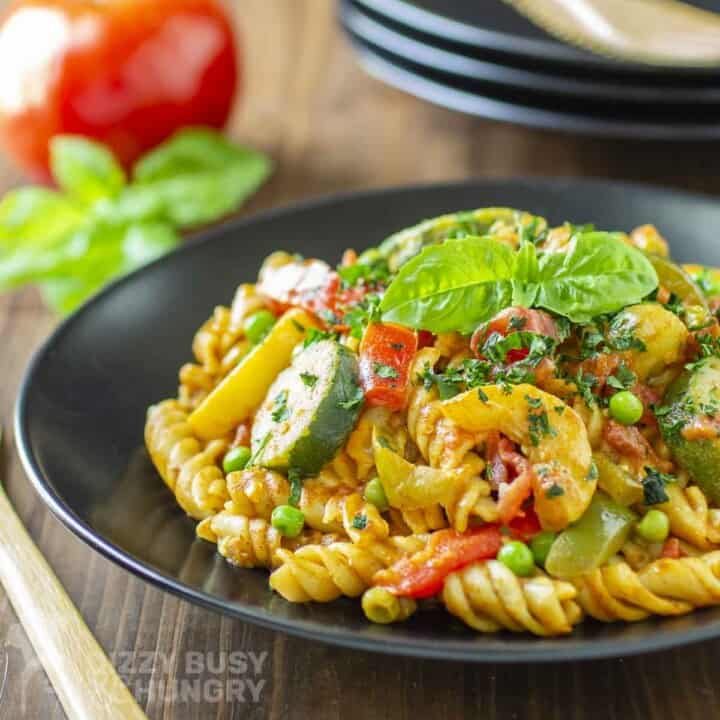 Side shot of pasta curry garnished with fresh basil in a black bowl with a tomato and basil in the background.