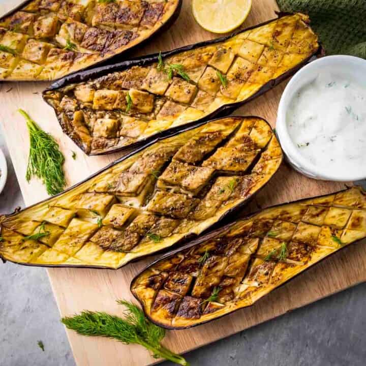 Overhead shot of four slices of roasted eggplant on a wooden cutting board with fresh dill, lemon slices, and dipping sauce on the side.