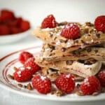Side shot of multiple waffle pieces stacked on each other with raspberries and pecans and topped with powdered sugar on a red and white plate.