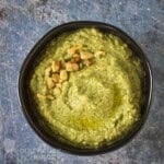 Overhead shot of zucchini pesto garnished with chopped walnuts in a black bowl on a blue marble surface.