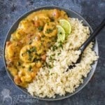 Overhead shot of coconut shrimp on one half of a black plate with rice on the other half garnished with fresh herbs and lime wedges with a black spoon on the side.
