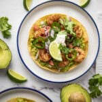 Overhead shot of two white and blue plates with shrimp tostadas garnished with fresh cilantro with lime wedges and avocados on the side.