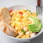 Tofu scramble in a white bowl served with extra tofu cubes, sliced cherry tomatoes, avocado wedges, and crusty bread.