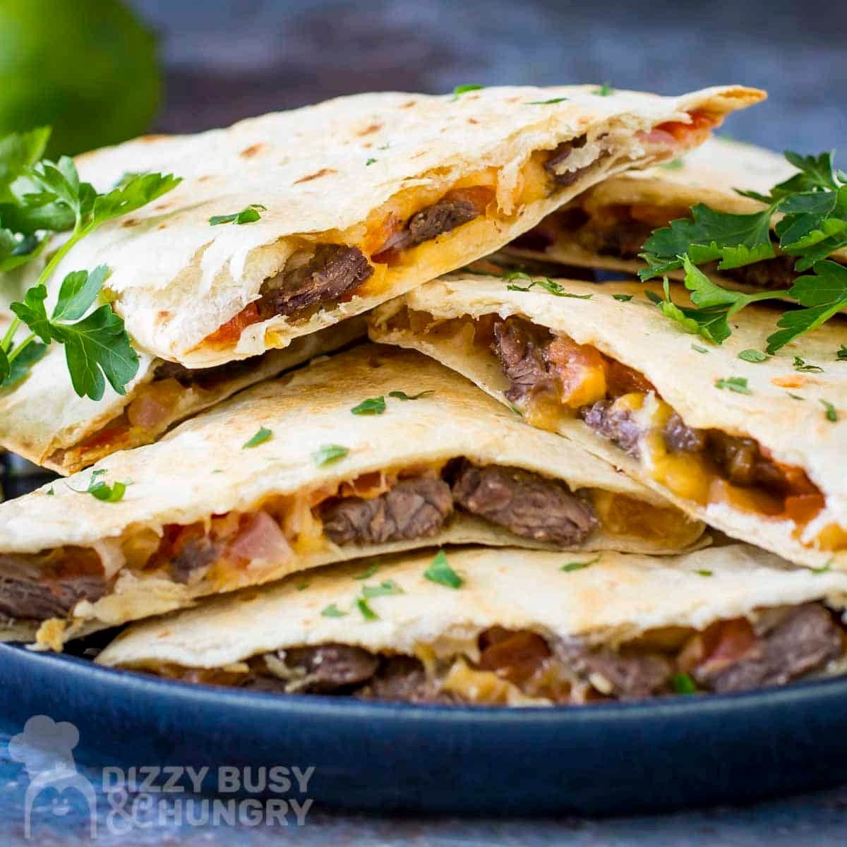 Side shot of multiple steak quesadillas stacked on a black plate with fresh herbs and lime wedges on the side.