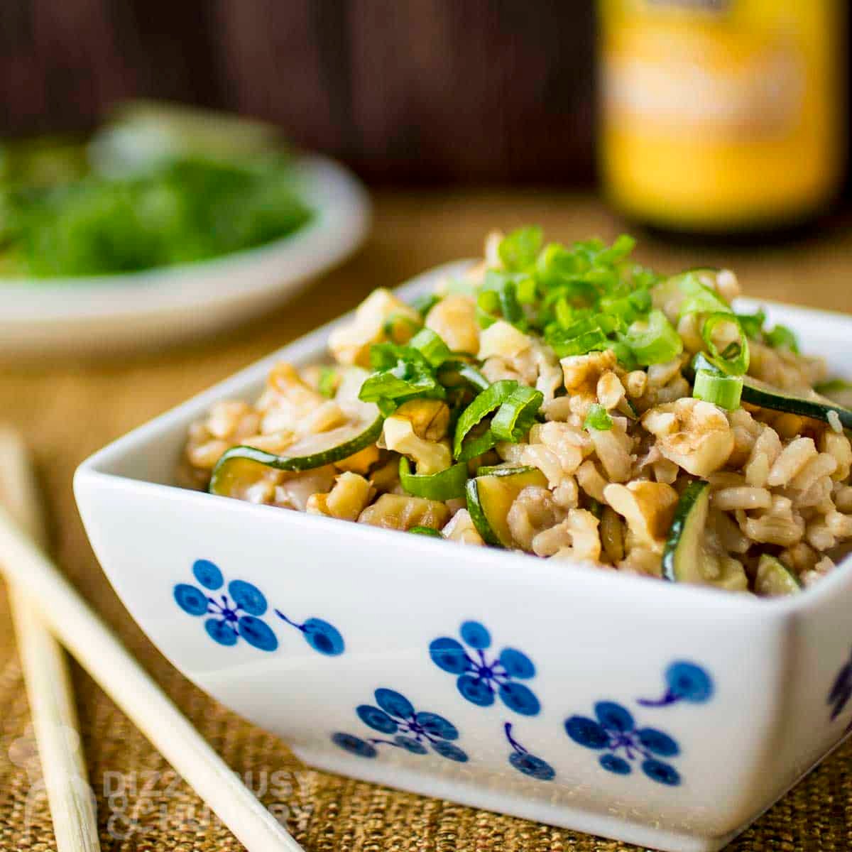 Side shot of a square white bowl with zucchini fried rice garnished with green onions on a burlap cloth with chopsticks on the side.