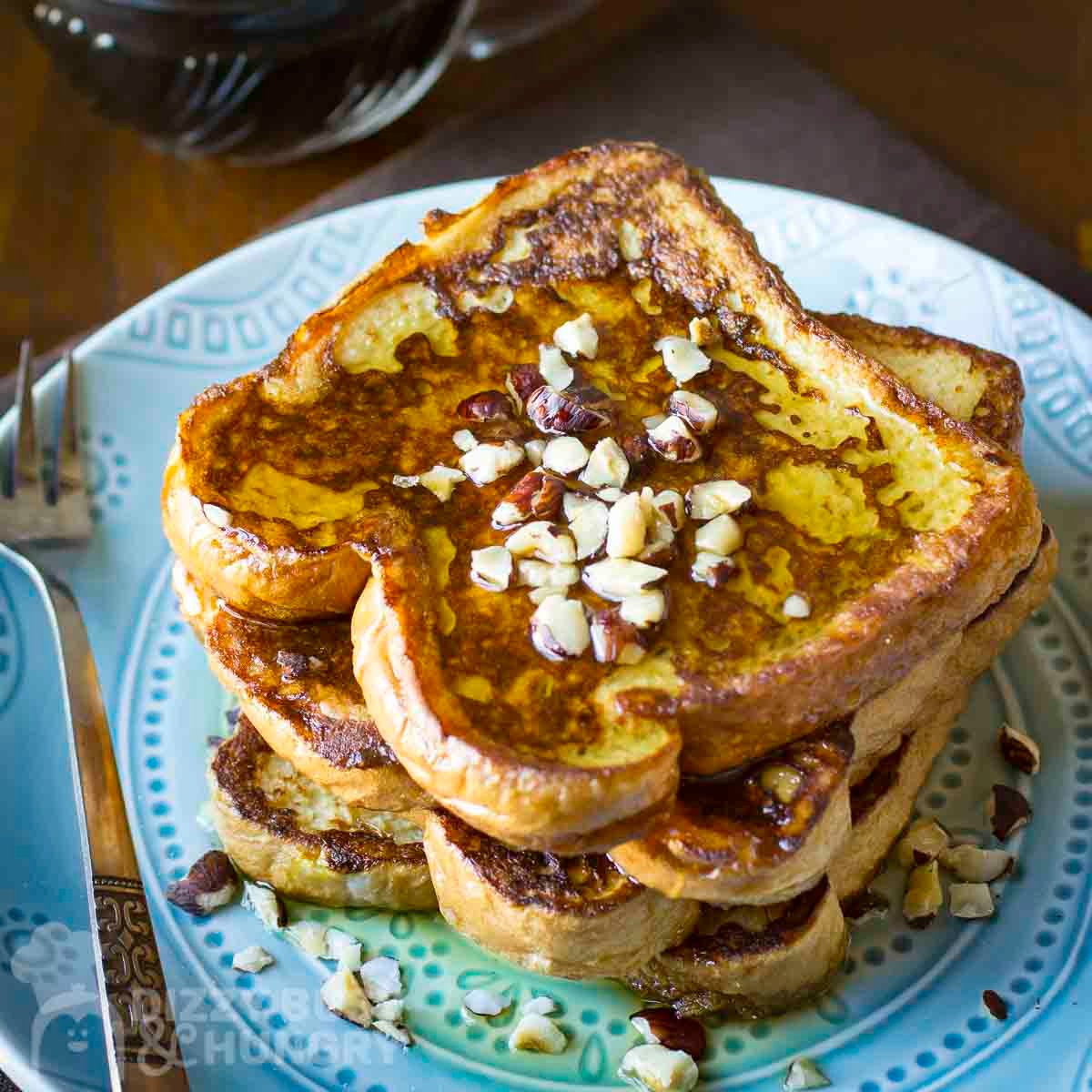 Side view of a stack of french toast with syrup and chopped almonds on a blue plate with a fork on the side.