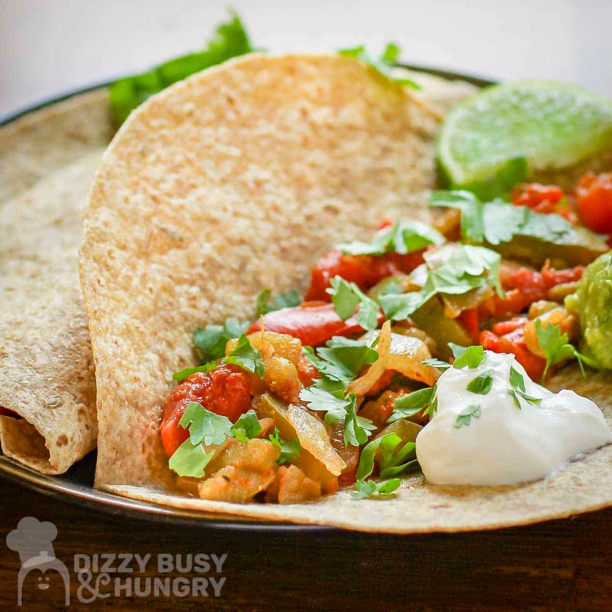 Close up shot of veggie fajitas in a tortilla garnished with cilantro and sour cream with a lime wedge on the side.