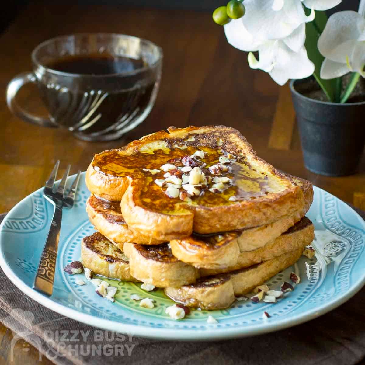 Side view of a stack of french toast with syrup and chopped almonds on a blue plate with a fork on the side and a cup of syrup and flowers in the background.