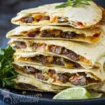 Side shot of multiple steak quesadillas stacked on a black plate with fresh herbs and lime wedges on the side.