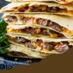 Side shot of multiple steak quesadillas stacked on a black plate with fresh herbs and lime wedges on the side.