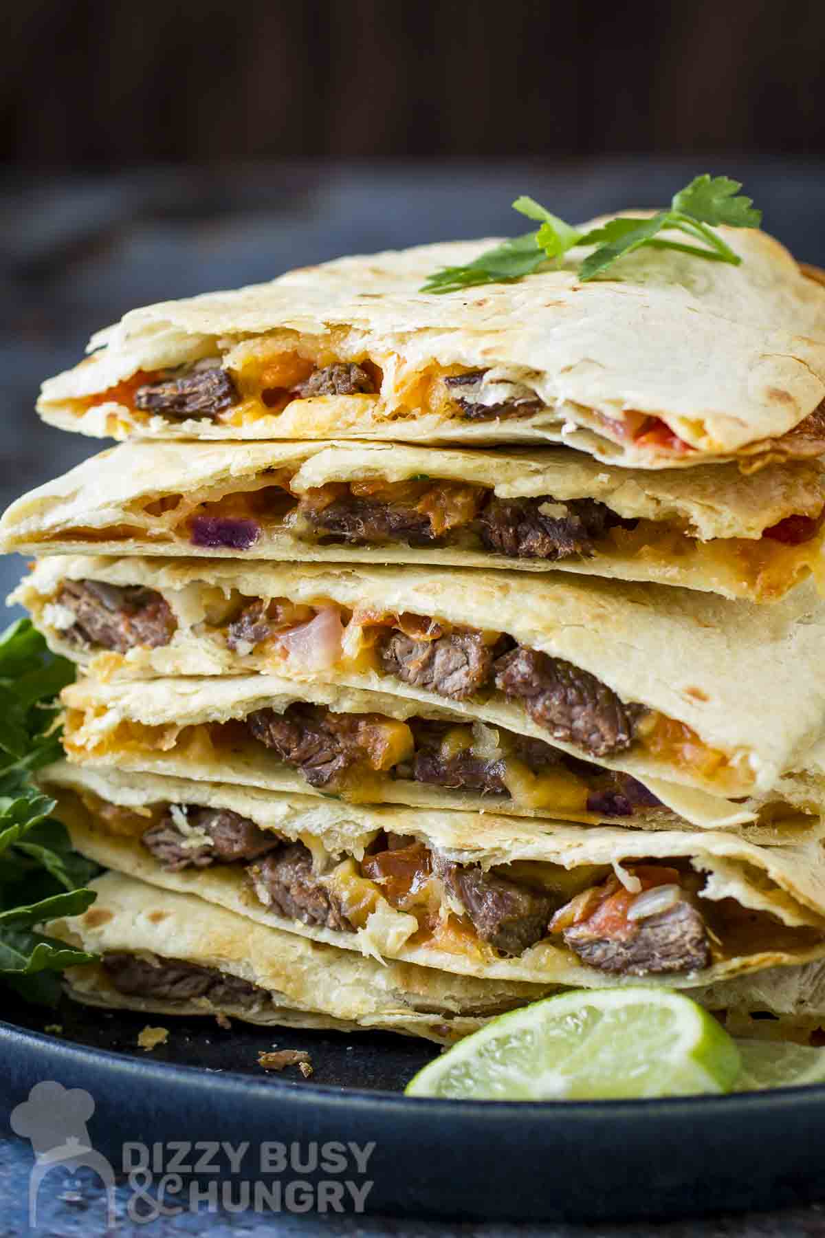 Side shot of multiple steak quesadillas stacked on a black plate with fresh herbs and lime wedges on the side.