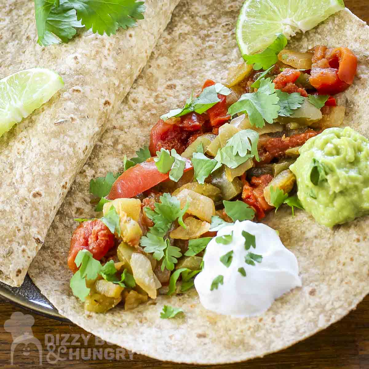 Overhead close up shot of veggie fajitas in a tortilla garnished with cilantro and sour cream with a lime wedge on the side.
