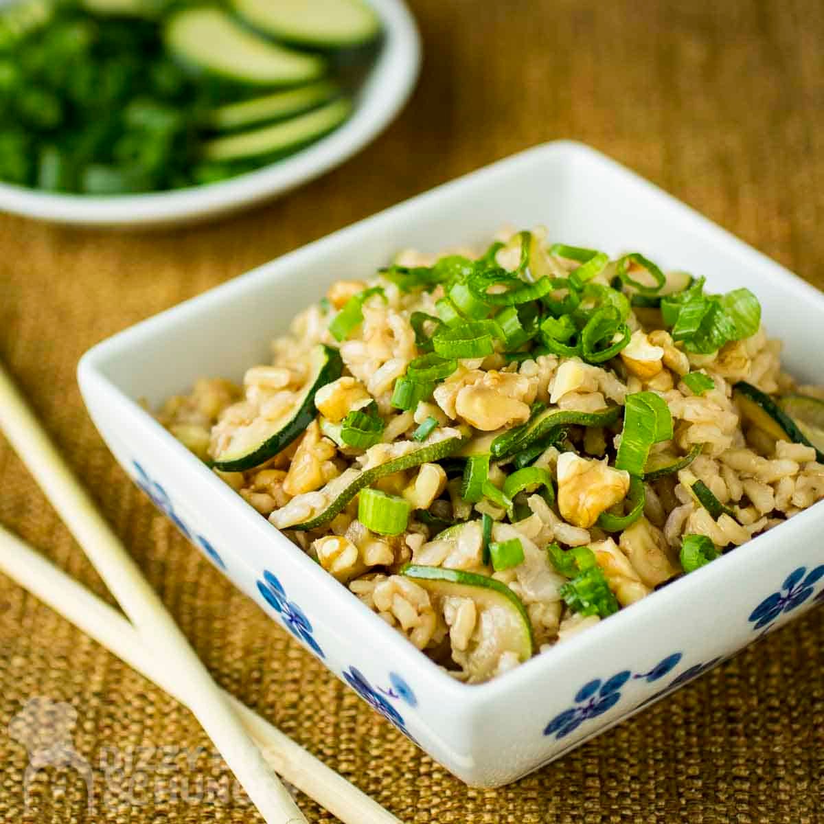 Side shot of a square white bowl with zucchini fried rice garnished with green onions on a burlap cloth with chopsticks on the side.