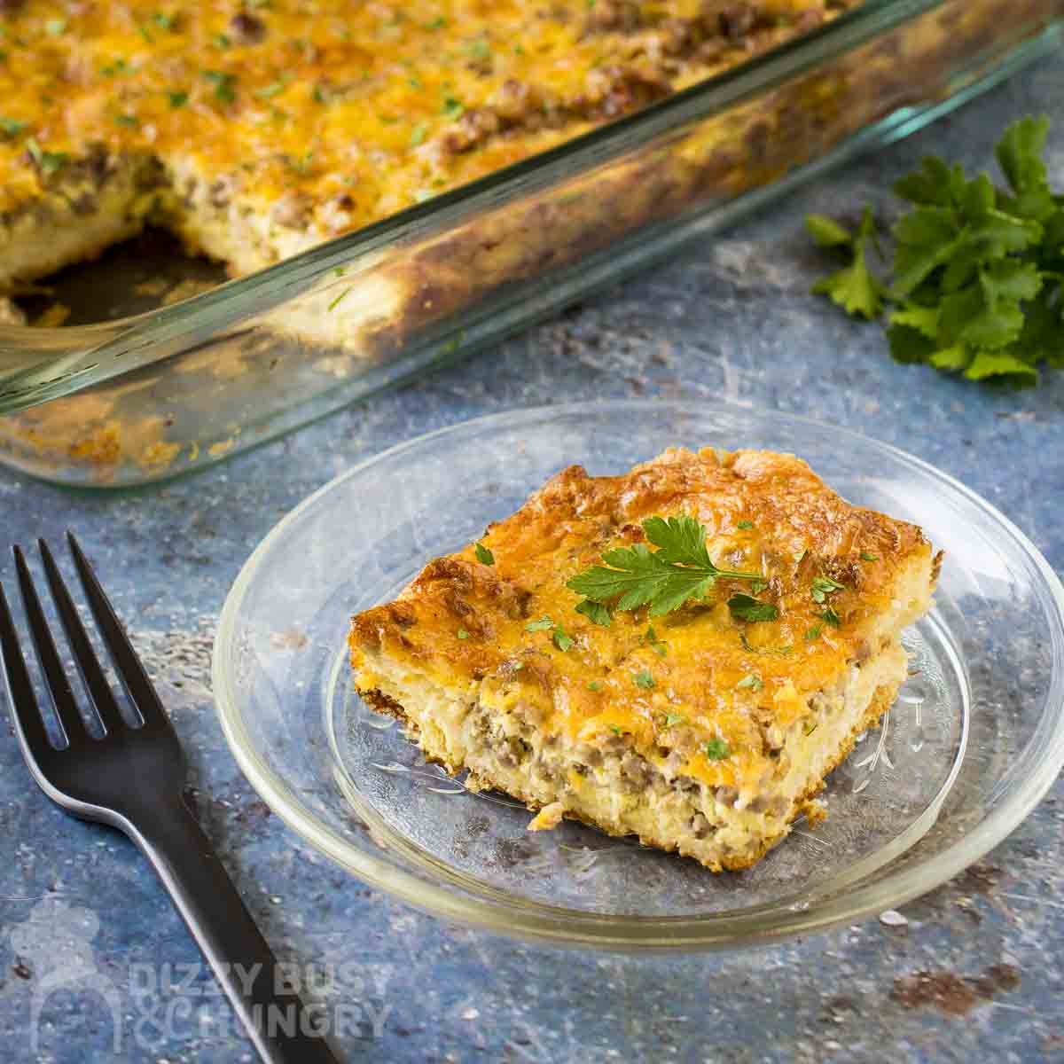 Side shot of a piece of crescent roll casserole on a clear plate with a black fork on the side with a full baking dish of the casserole and fresh herbs in the background.