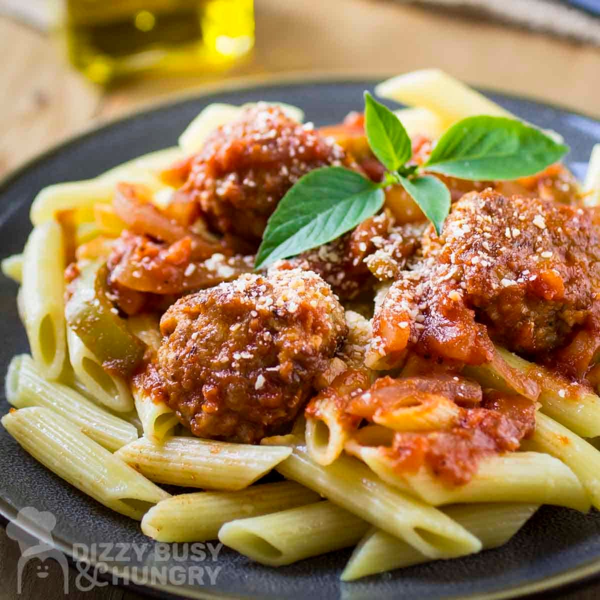 Side shot of meatballs and sauce over a bed of penne garnished with fresh basil on a black plate.