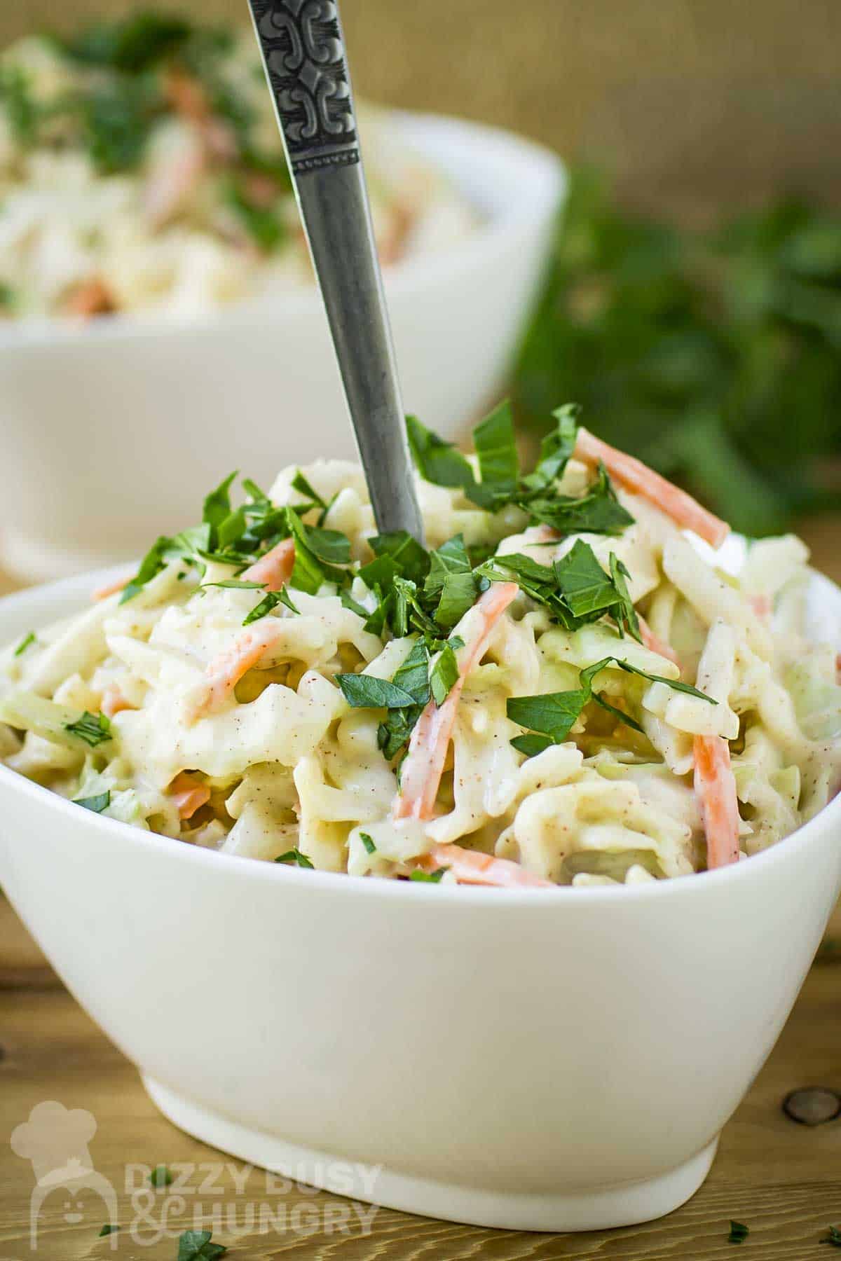 Side shot of a white bowl with chipotle coleslaw garnished with fresh herbs on a wooden table with more herbs and another bowl in the background.