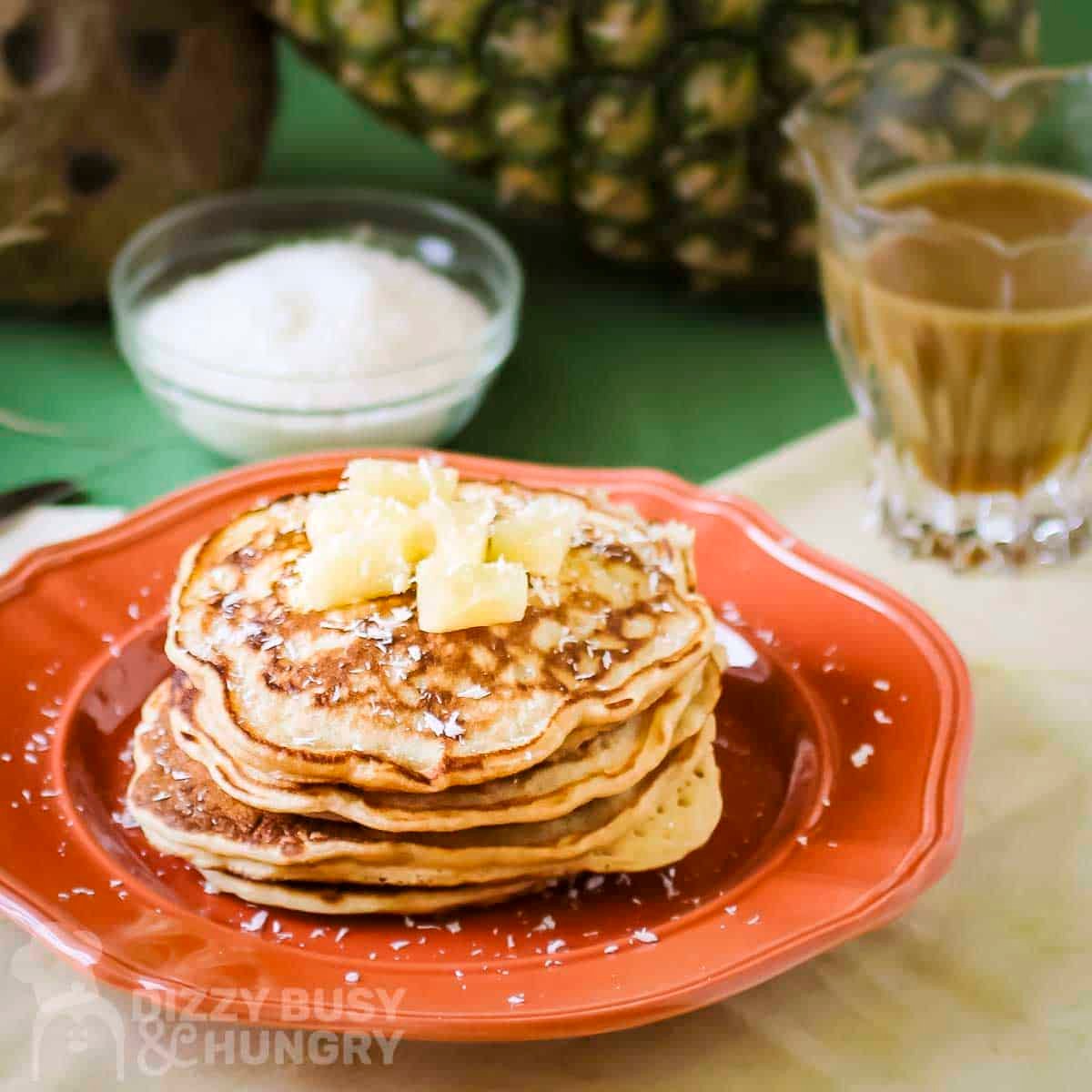 Side shot of a stack of pineapple pancakes garnished with coconut shreds and pineapple chunks on an orange plate.