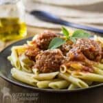 Side shot of meatballs and sauce over a bed of penne garnished with fresh basil on a black plate.