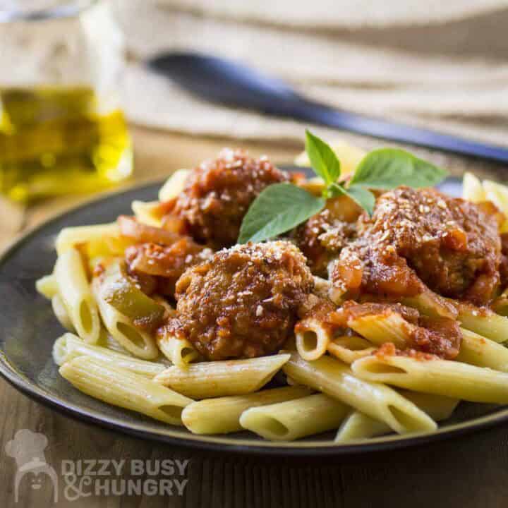 Side shot of meatballs and sauce over a bed of penne garnished with fresh basil on a black plate.