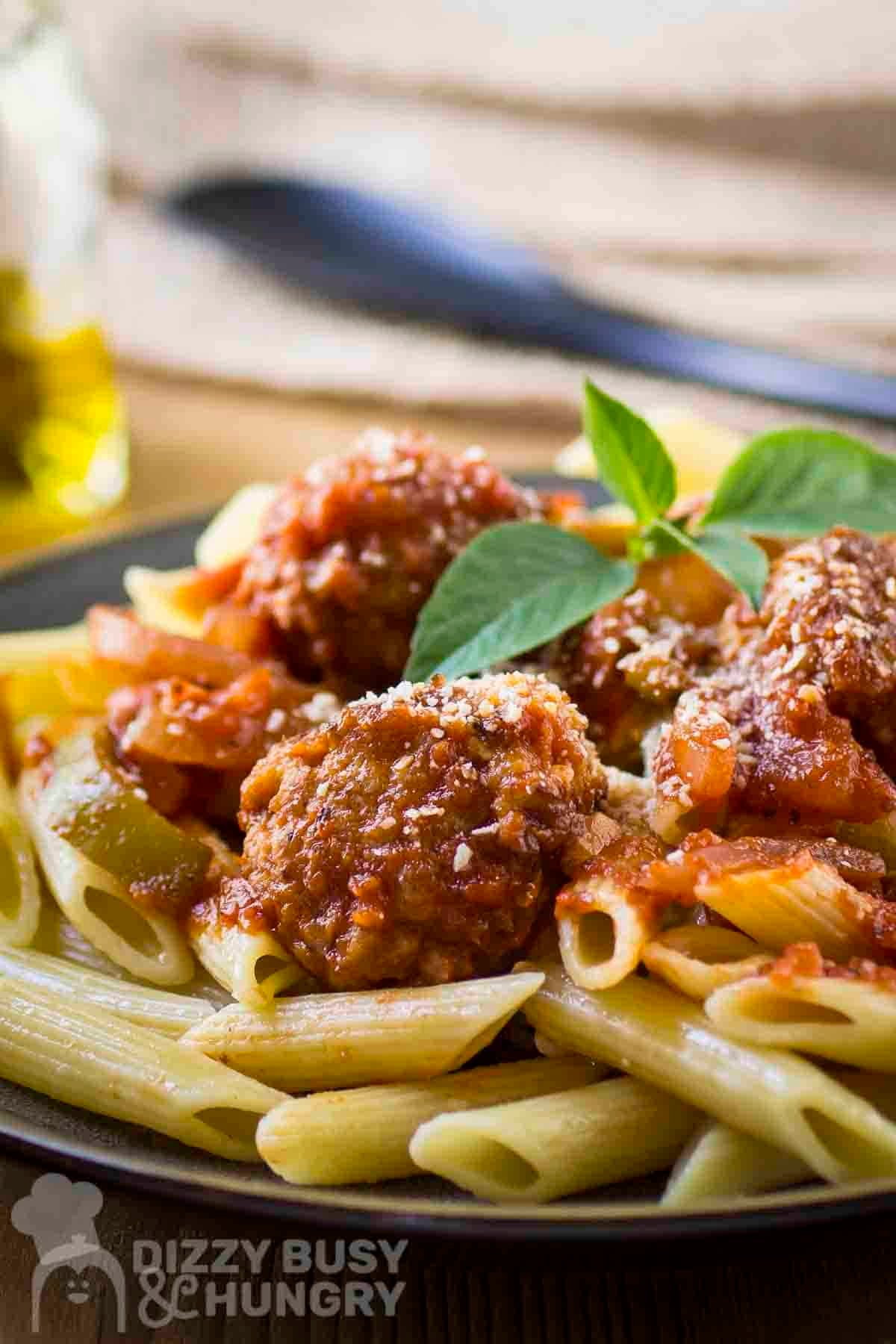Side shot of meatballs and sauce over a bed of penne garnished with fresh basil on a black plate.