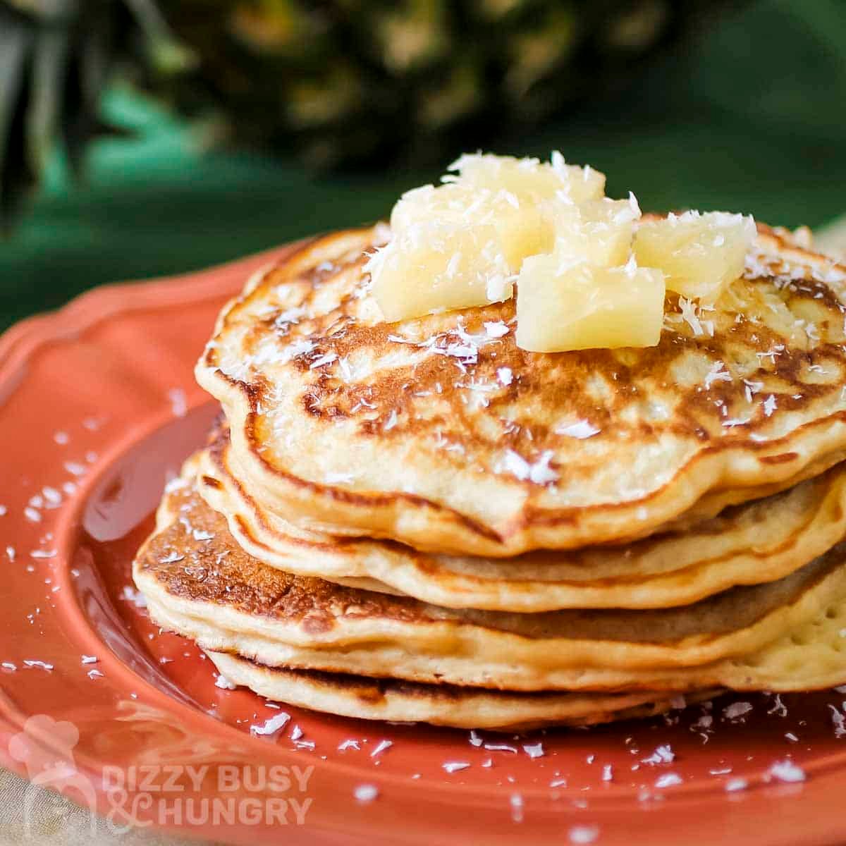 Side shot of a stack of pineapple pancakes garnished with coconut shreds and pineapple chunks on an orange plate.