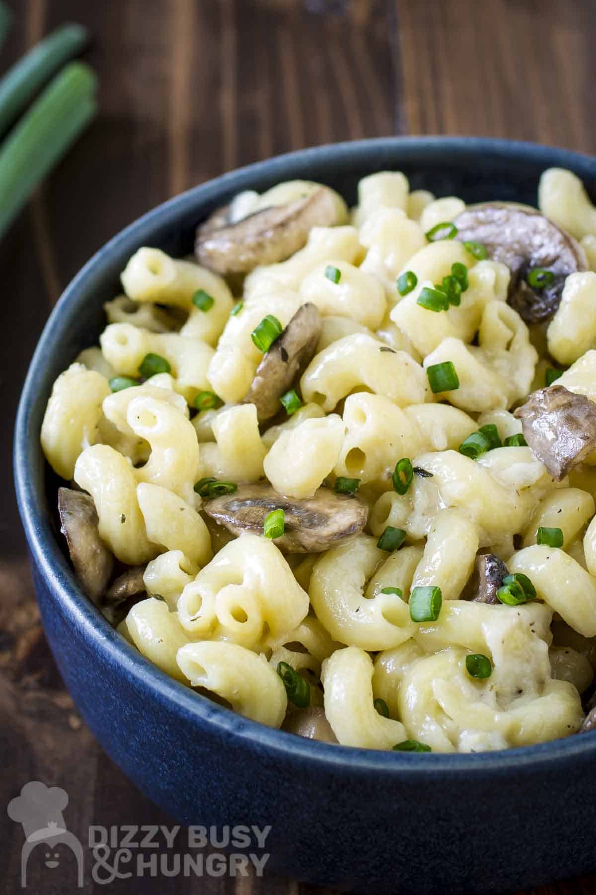 Side shot of mushroom mac and cheese garnished with fresh green onions slices in a blue bowl with more green onions on the side.