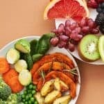 Colorful snack plates on a tangerine-colored background with grapefruit, berries, grapes, kiwi, apples, sweet potatoes, veggies, and beans.