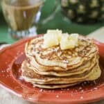 Side shot of a stack of pineapple pancakes garnished with coconut shreds and pineapple chunks on an orange plate.